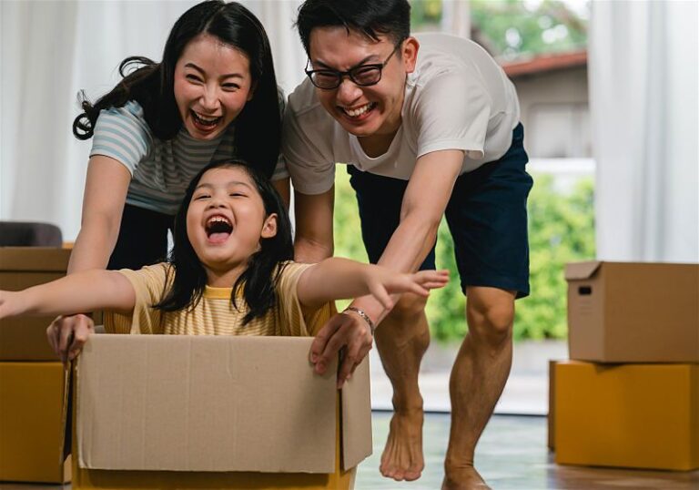 Menina sentada em caixa de papelão com pai e mãe ajudando a empurrar, todos sorridentes